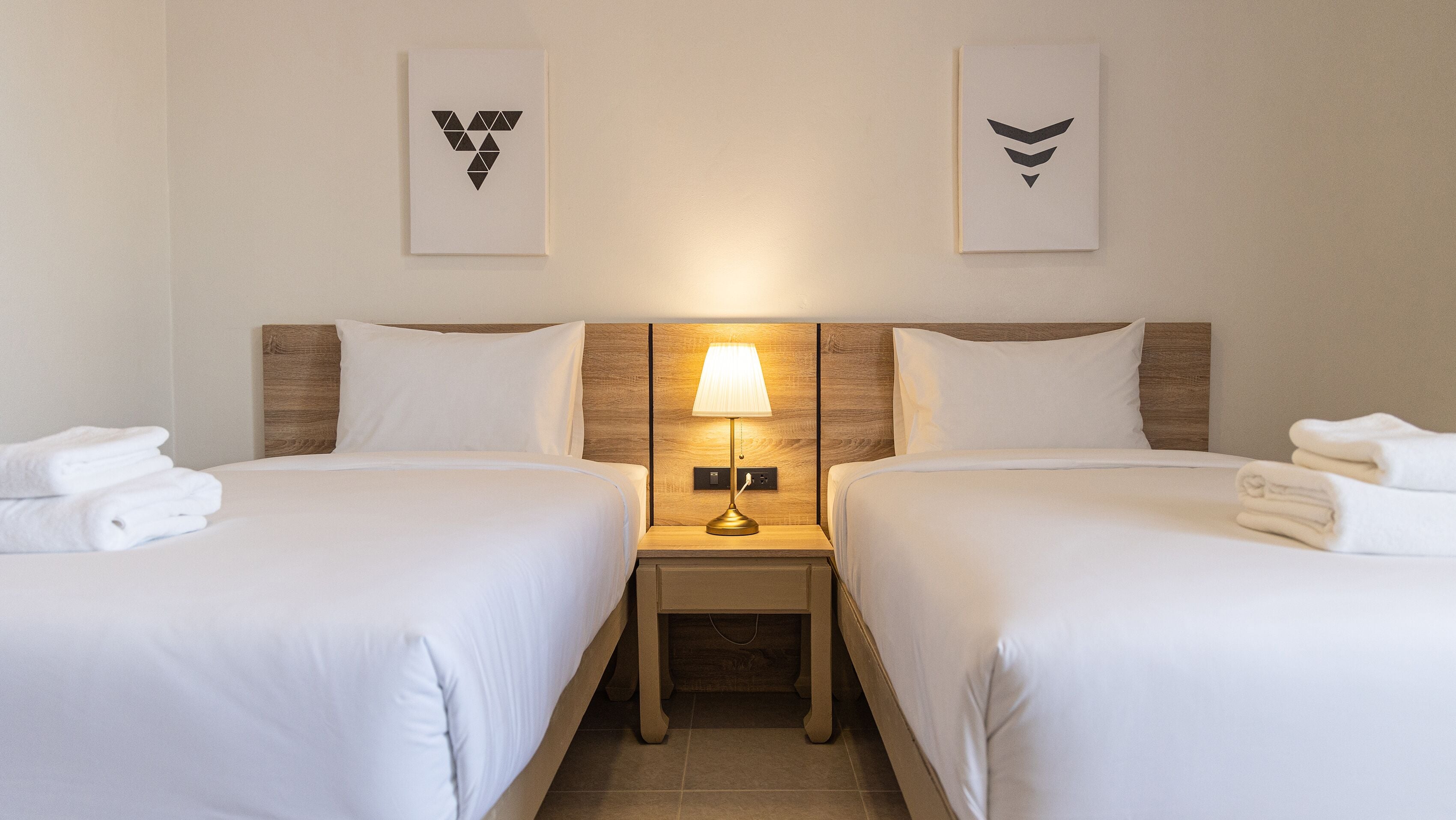 A hotel room with two beds, a nightstand with a lamp, and artwork on the walls.