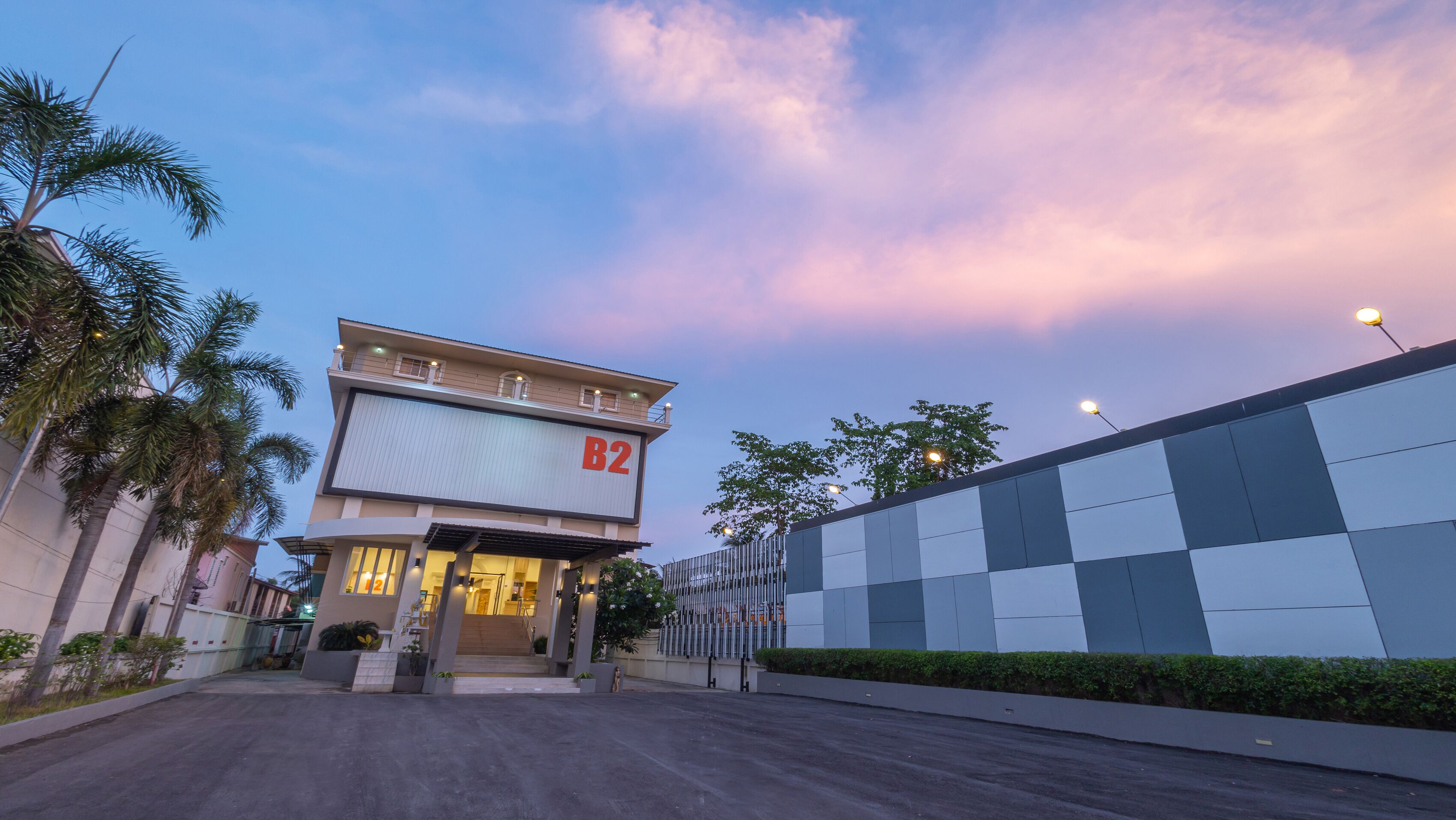 A modern building with a sign 'B2' at the entrance, surrounded by palm trees and a gated area.