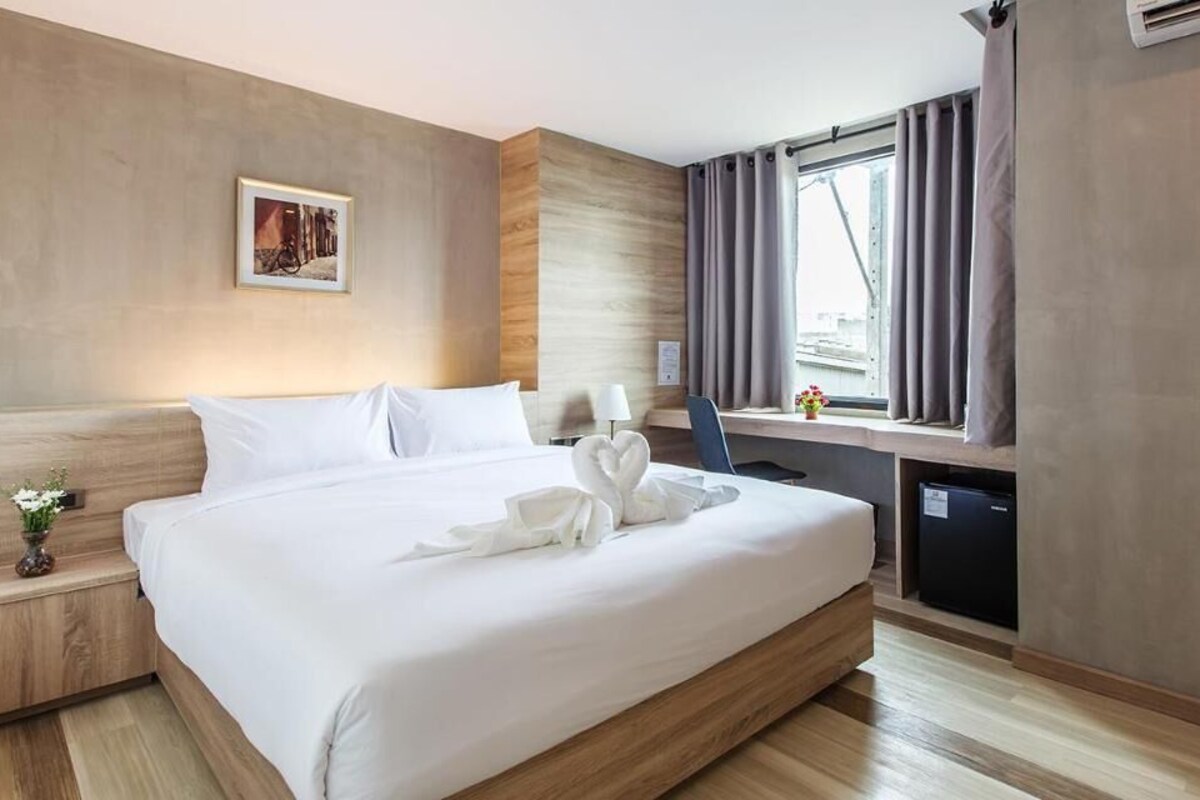 A hotel room with a large bed, a desk, a chair, a small table with flowers, and a window with curtains.
