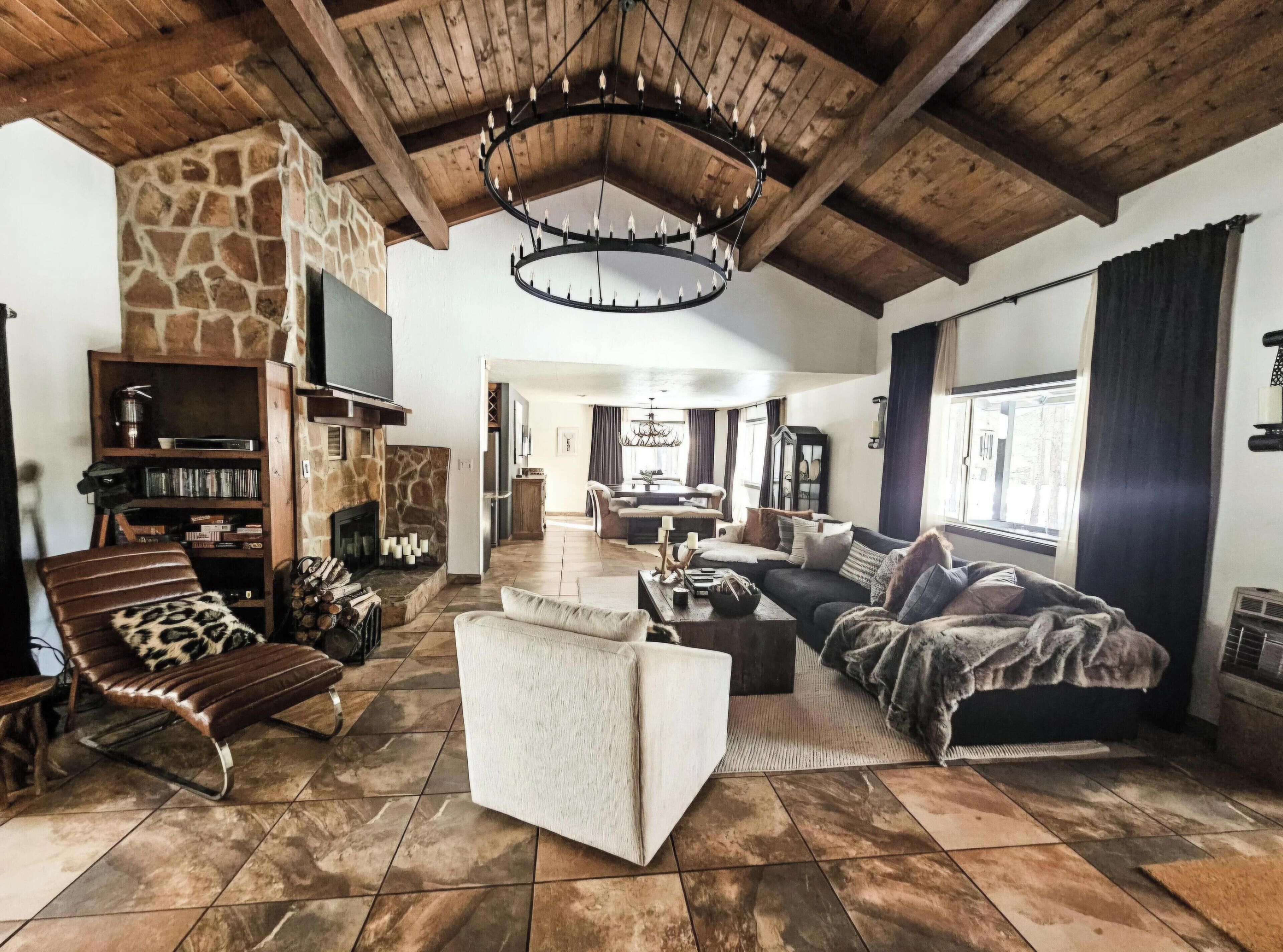 A spacious living room with a stone fireplace, a wooden ceiling, and a chandelier. There is a leather chair, a sofa, and a coffee table.