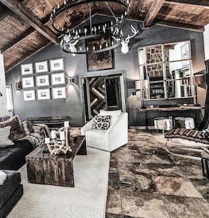 A rustic interior with a high ceiling, a chandelier, a fireplace, a sofa, a coffee table, and a bookshelf. - Luxury Angel Fire Retreat - Fireplace, Skiing & Mountain Charm (Angel Fire)