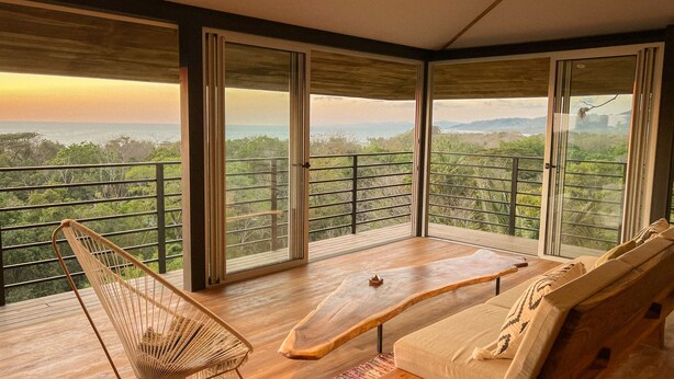 Un ampio balcone con pavimento in legno, una sedia in vimini, un divano e un tavolo.