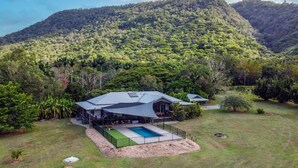 Une maison moderne dotée d’une grande terrasse, d’une piscine et d’un jardin luxuriant, le tout entouré d’une forêt dense et de collines.