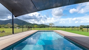 A rectangular swimming pool with a clear blue water surface, surrounded by a black metal fence, under a shaded area with a view of a green landscape and mountains in the background.