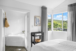 A hotel room with a large bed, a bathtub, a small shelf with a bowl, a painting, and a view of greenery through the window.