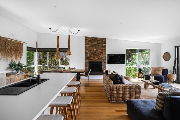 A modern kitchen with a central island, a dining area with wooden chairs, a living room with a sofa and a fireplace, and large windows offering a view of greenery.