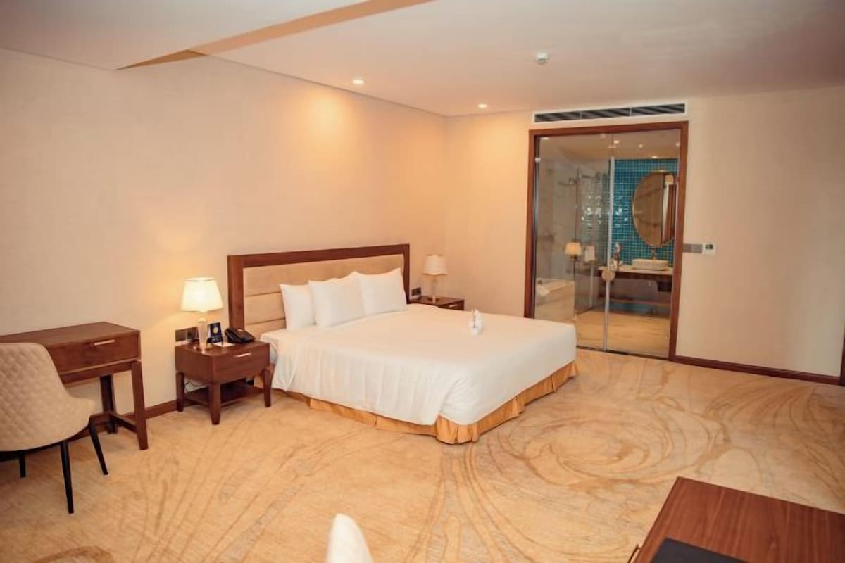 Ein Hotelzimmer mit einem großen Bett, einem Schreibtisch, einem Stuhl und einem durch eine Glastür einsehbaren Badezimmer.