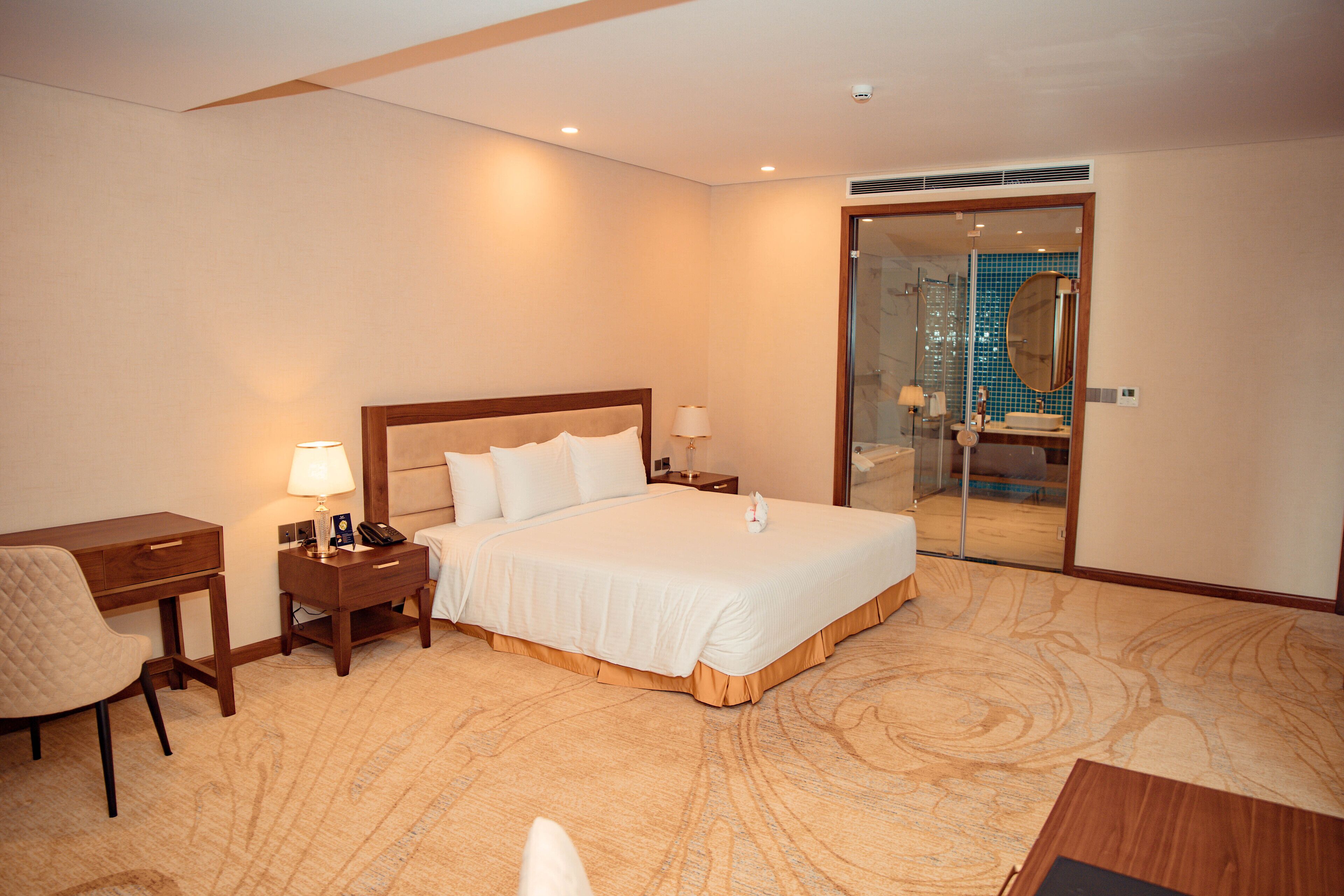 A hotel room with a large bed, a desk, a chair, and a bathroom visible through a glass door.