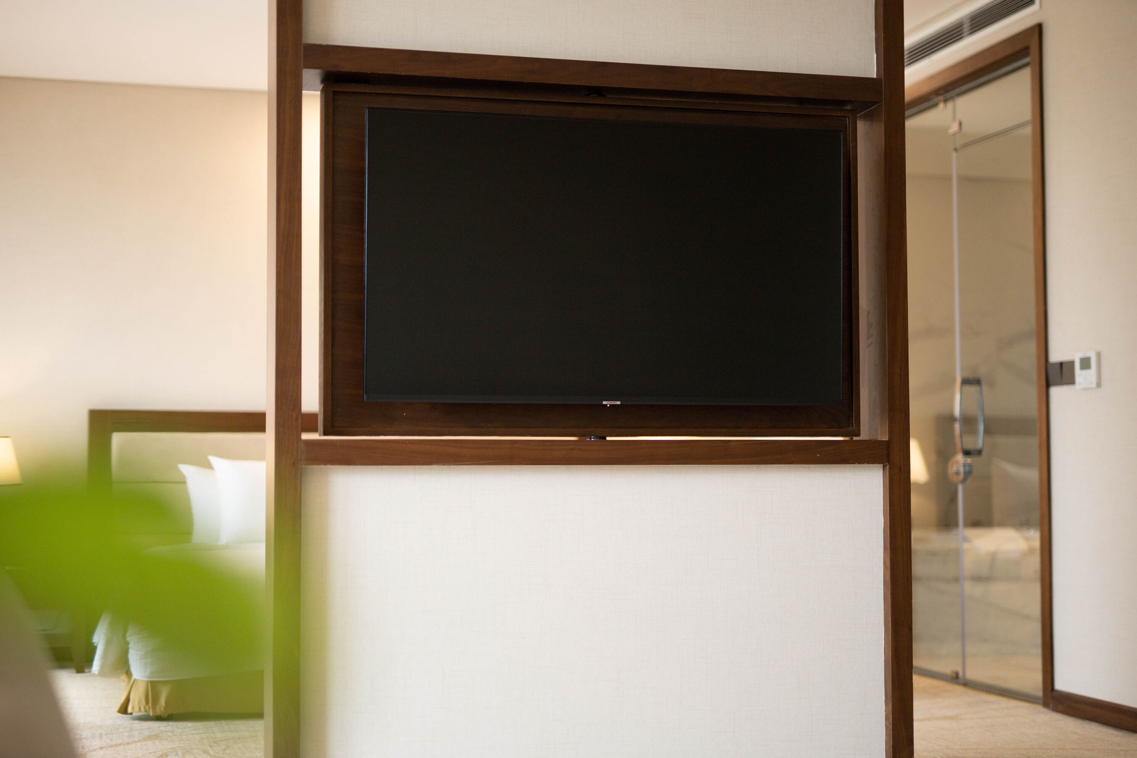 A hotel room with a large flat-screen TV mounted on a wall, a bed with white bedding, and a glass shower door.