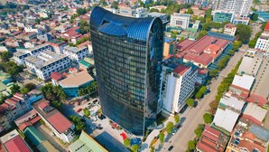 A modern high-rise building with a curved glass facade, surrounded by a dense urban area with various buildings and streets. - Muong Thanh Luxury Cao Bang Hotel (Thuc Phan)