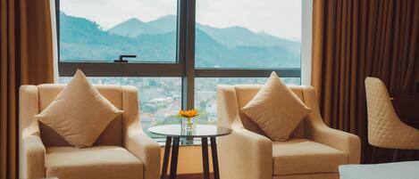 A room with two beige armchairs, a small round table with a vase of flowers, and a large window offering a mountain view.