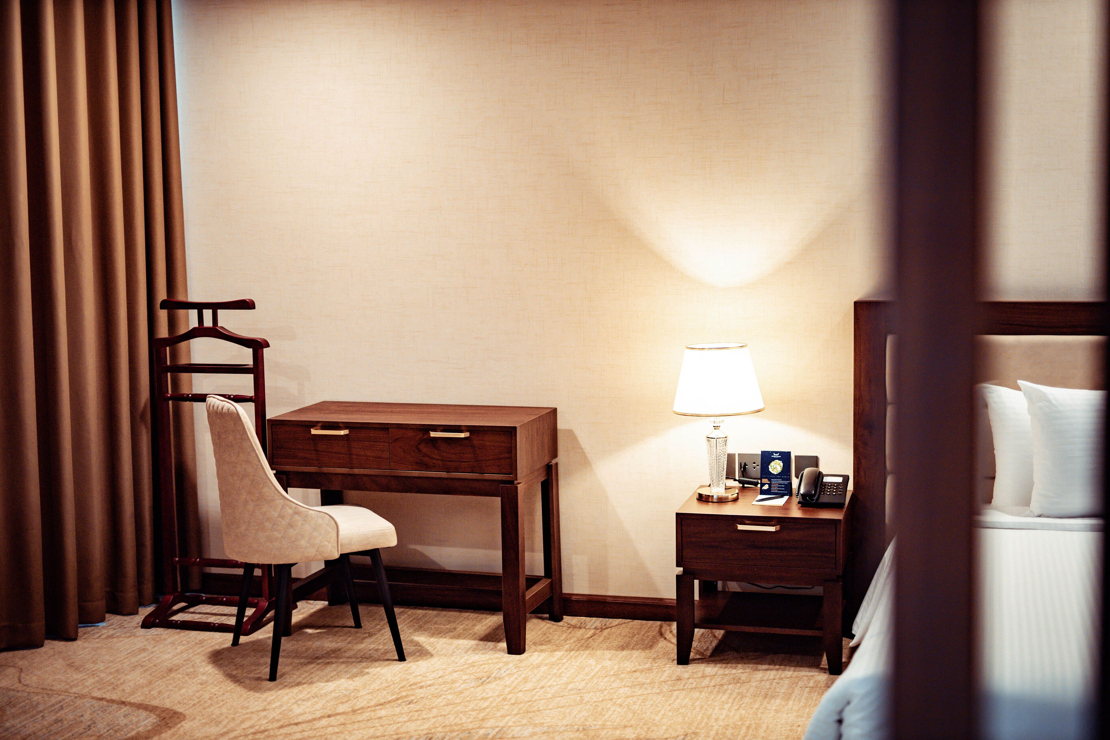 A hotel room with a wooden desk, a chair, a bedside table with a lamp, and a bed.