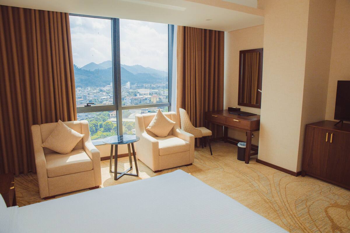 Quarto de hotel com duas poltronas, uma escrivaninha, uma cama grande e vista para a cidade através de uma janela ampla.