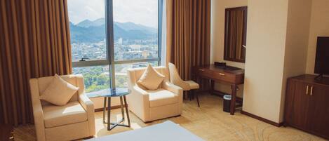 Camera d'albergo con due poltrone, una scrivania, un letto grande e vista sulla città attraverso un'ampia finestra.