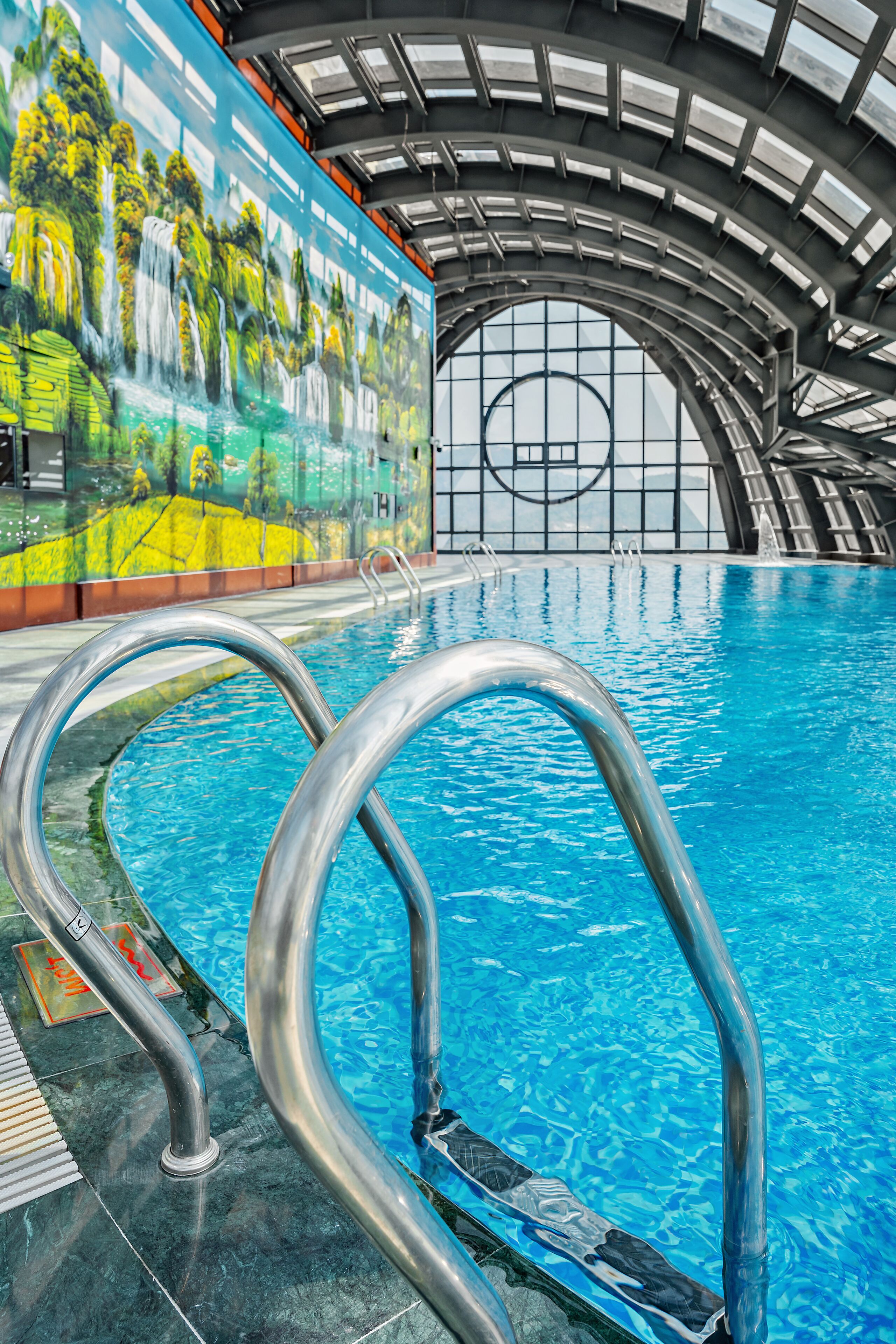 Una piscina interior con agua cristalina, escaleras metálicas y un mural de cascada en la pared.