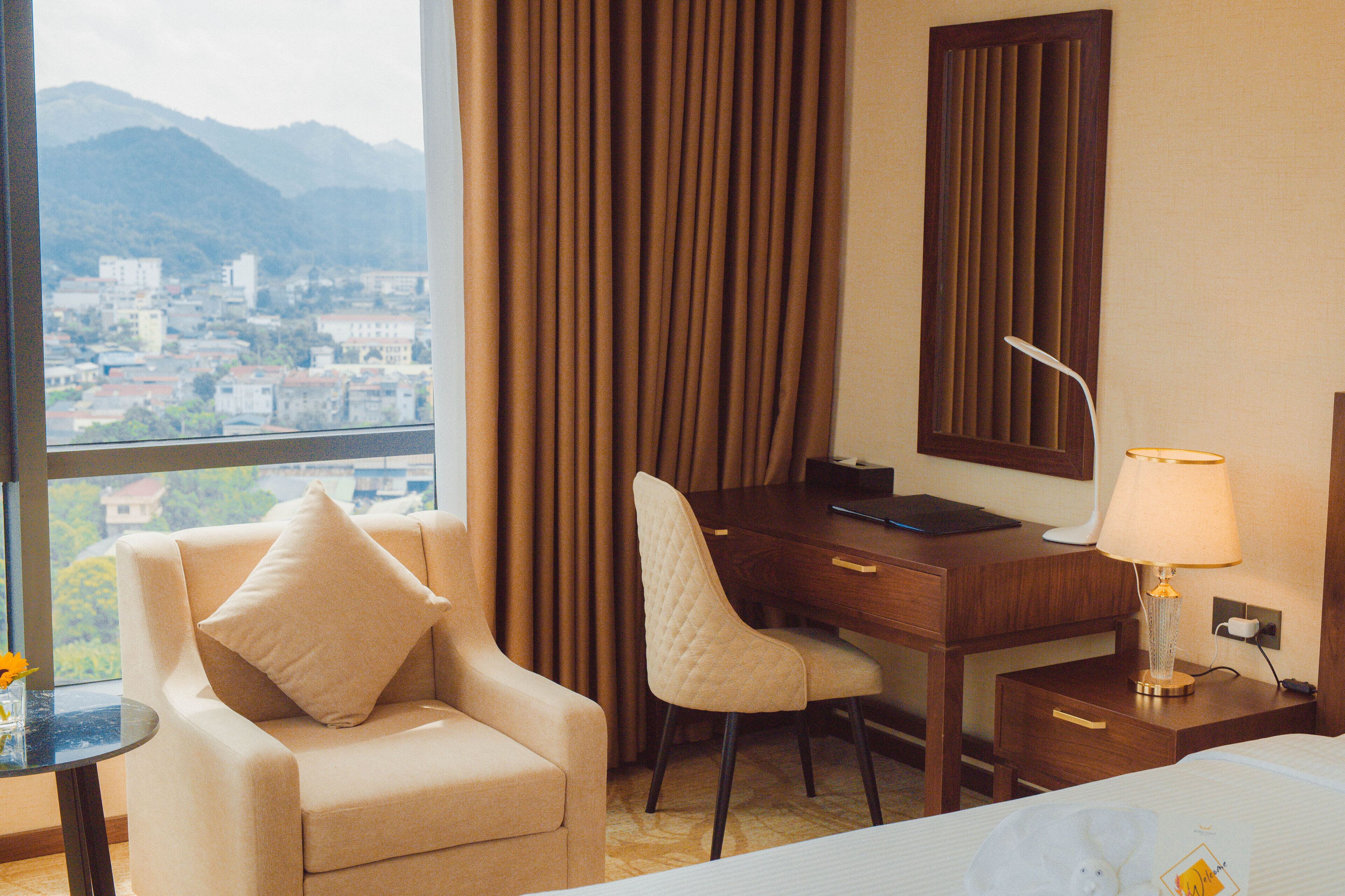 Habitación de hotel con cama, escritorio con silla, lámpara y vista a un paisaje urbano a través de una ventana.