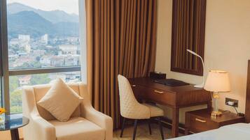 Habitación de hotel con cama, escritorio con silla, lámpara y vista a un paisaje urbano a través de una ventana.