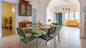 Ein Essbereich mit einem Tisch für vier Personen, umgeben von Stühlen, einem Schrank mit Glastüren und Blick auf ein gelbes Sofa durch einen Rundbogen.