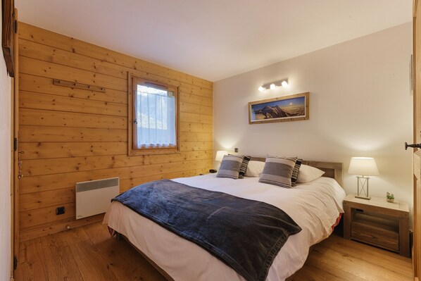 Une chambre avec un mur en bois, un lit recouvert d’une couverture sombre, deux lampes de chevet, une table de nuit et un tableau accroché au mur.