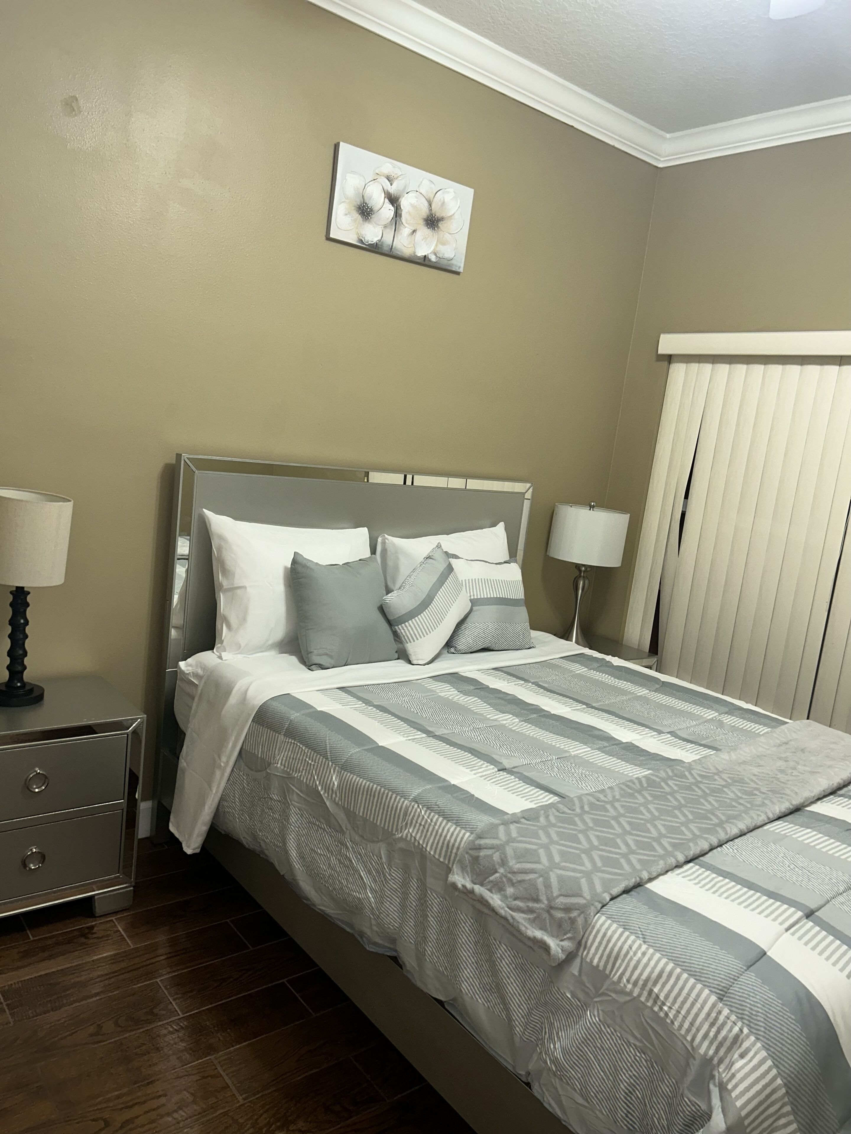 A bedroom with a bed, two bedside tables, a nightstand, a window with blinds, and a wall-mounted artwork.