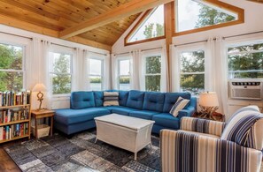 A cozy living room with a blue sectional sofa, a wicker ottoman, striped armchairs, a bookshelf, and large windows offering a view of trees. - Quaint lakefront; firepit, boat, kayaks, paddleboard, grill, laundry, hammock (Springfield)