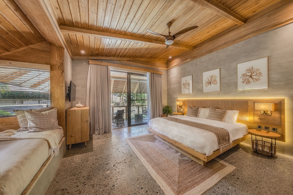 Une chambre spacieuse avec un grand lit, un lit plus petit, un ventilateur de plafond et une vue sur l’extérieur.