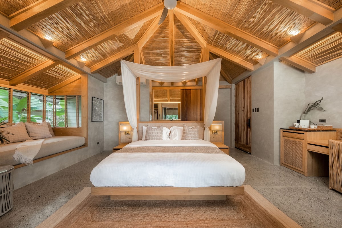 Une chambre spacieuse avec un grand lit, des poutres apparentes en bois au plafond et une vue sur la verdure environnante.