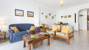 Ein Wohnzimmer mit einem blauen Sofa, einem Holztisch und zwei gemusterten Sesseln. An den Wänden befinden sich dekorative Wandteller und gerahmte Bilder.