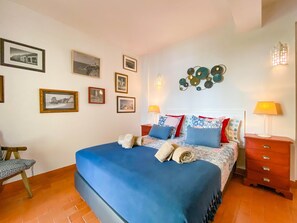Ein Schlafzimmer mit einem großen Bett, vielen Kissen, einem Nachttisch, einem Sessel und mehreren gerahmten Bildern an der Wand.
