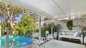 A covered outdoor area with a swimming pool, white outdoor furniture, and lush greenery.