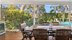 An outdoor patio with a wooden table and chairs, surrounded by lush greenery and a clear view of a swimming pool.