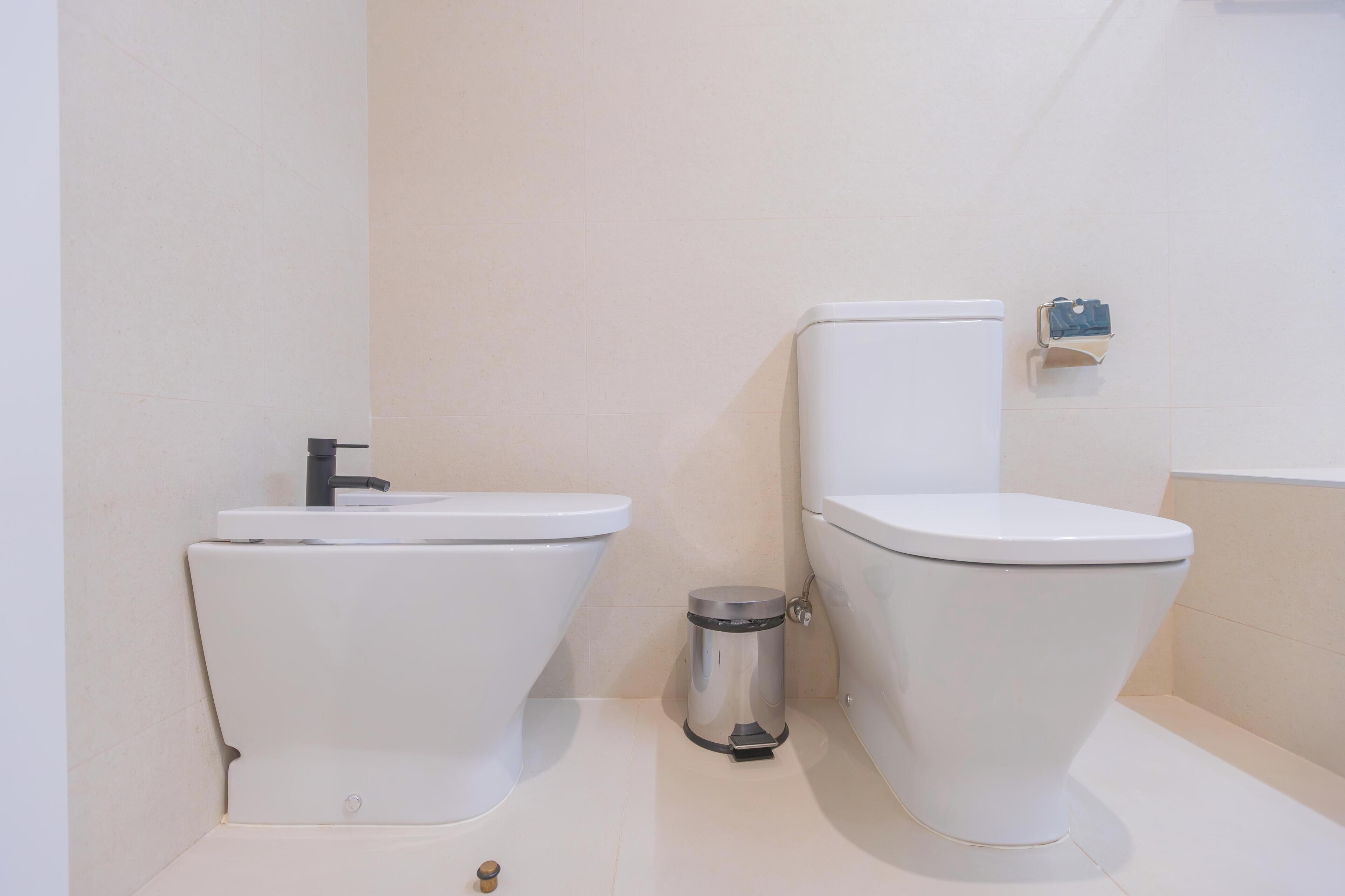 A bathroom with two toilets, a sink, and a trash bin.
