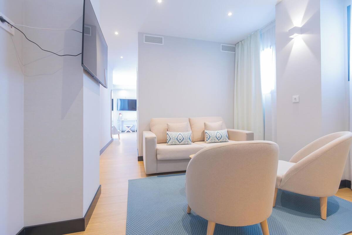 Ein modernes Wohnzimmer mit einer Couch und zwei Sesseln, einem flachbildfernseher an der Wand und einem Fenster mit Vorhängen.