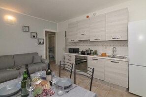 A modern kitchen with light wood cabinets, a dining area with a table set for two, and a grey sofa in the living area.