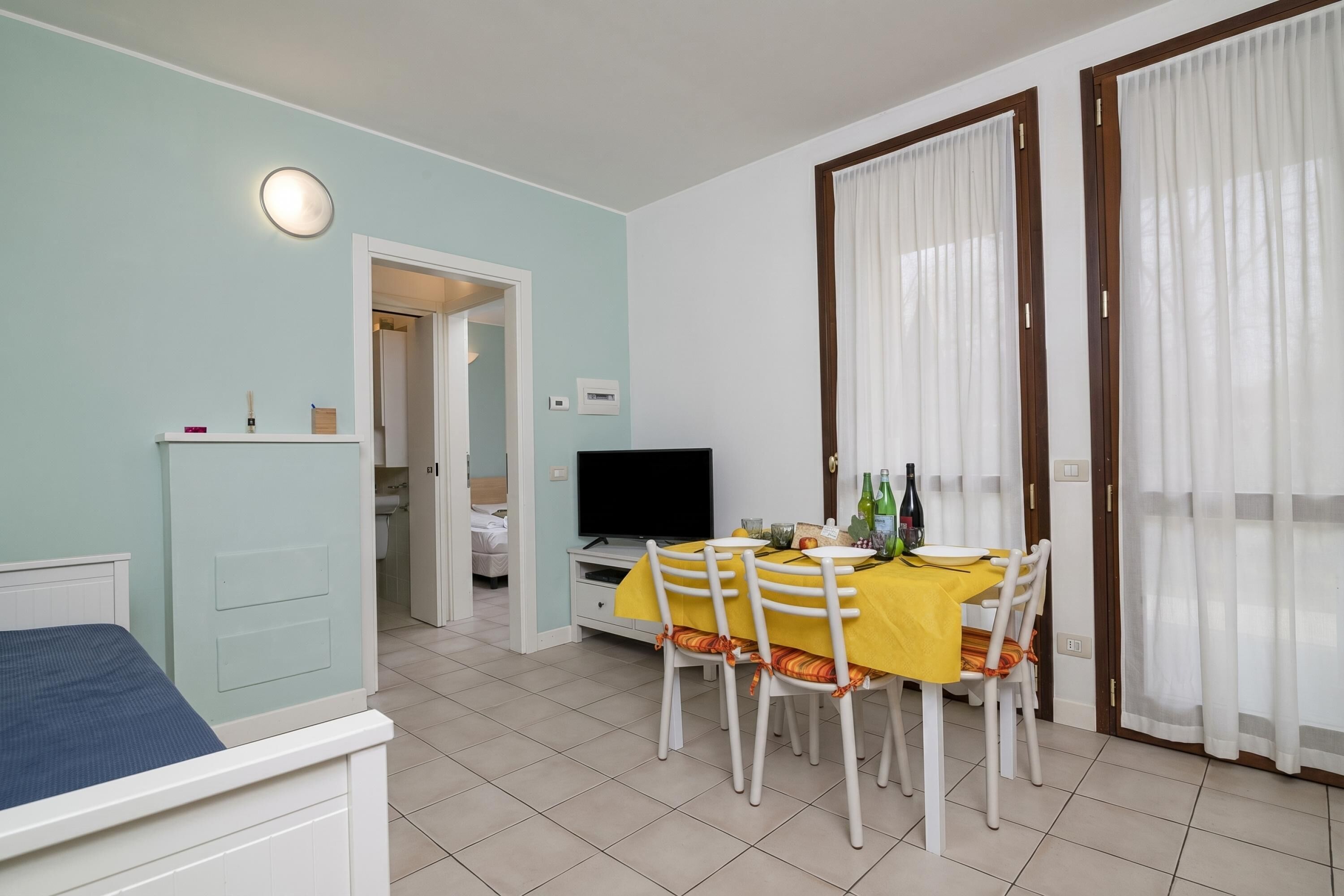A room with a bed, a dining table with chairs, a TV, and a door leading to another room.