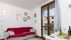 A living room with a red sofa, a dining table with chairs, a window with curtains, and a view of buildings outside. - Porto Letizia Torino 1B, Porlezza, Italy (Porlezza)