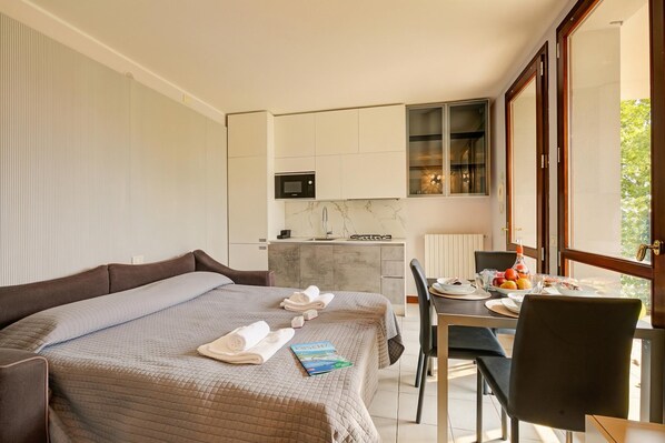A hotel room with a bed, a dining table with chairs, a kitchen area, and a large window.