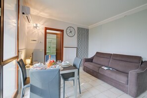 A living room with a brown sofa, a dining table with chairs, a clock on the wall, and a patterned curtain.
