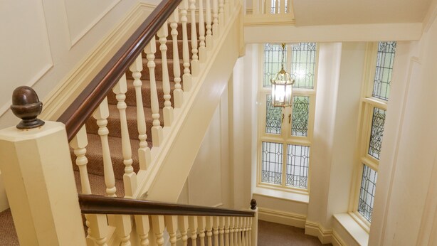 Eine imposante Treppe mit hölzernem Geländer und weißen Spindeln, die zu einer Zwischenebene mit einem großen Fenster mit verzierten Glasscheiben führt.