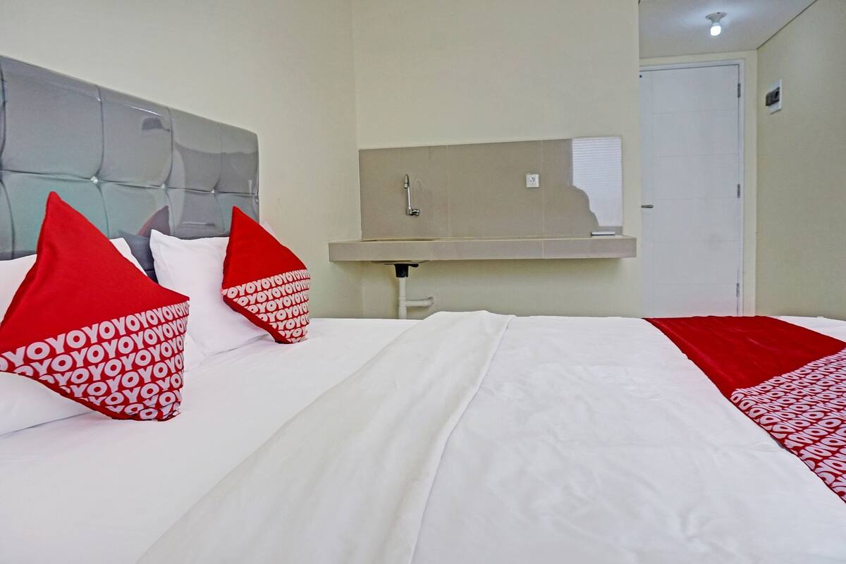 A hotel room with a bed, red and white pillows, a headboard, a mirror, a sink, and a door.