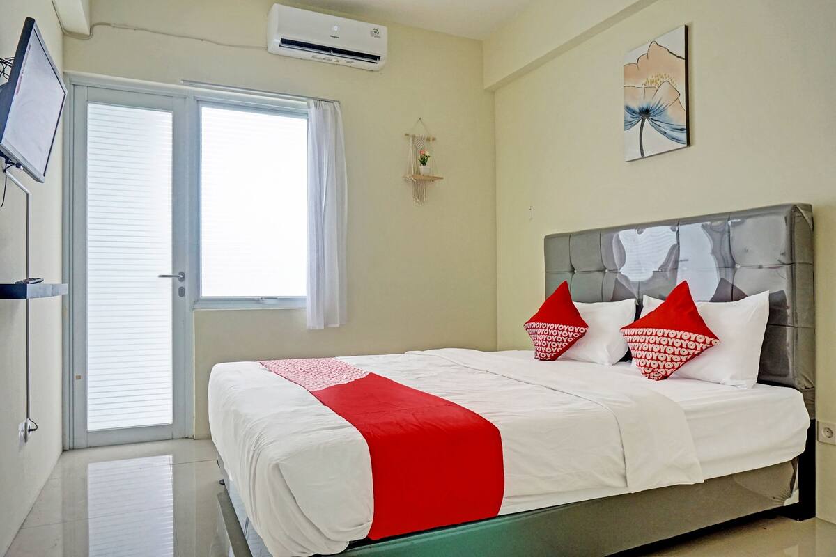 A hotel room with a bed, a TV, an air conditioner, a window with curtains, and a wall-mounted shelf with a plant.