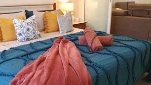 Una cama con colcha azul y toallas rosas, cabecera de madera y mesita de noche con lámpara.