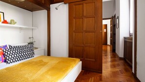 A bedroom with a bed, a wooden wardrobe, a white shelf with a plant, and a hallway with a wooden floor. - GuestHost - Spacious two-room apartment with kitchen for 5 persons with one bedroom, living room with sofa bed and a loft (overlooking the rooms below) with a bed and office area. The kitchen/dining room is separate. In total 65 sqm in old Milan building (Milan)