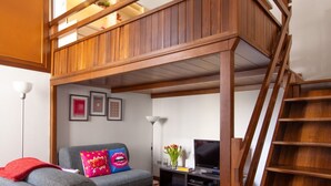 A loft-style living room with a wooden staircase, grey sofa, and a flat-screen TV. - GuestHost - Spacious two-room apartment with kitchen for 5 persons with one bedroom, living room with sofa bed and a loft (overlooking the rooms below) with a bed and office area. The kitchen/dining room is separate. In total 65 sqm in old Milan building (Milan)