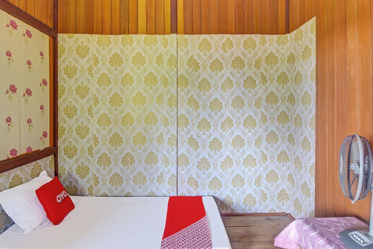 A room with a bed, a red pillow, a fan, and patterned wallpaper.