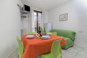 A dining area with a table set for two, a green sofa, a window with curtains, and a wall-mounted TV.