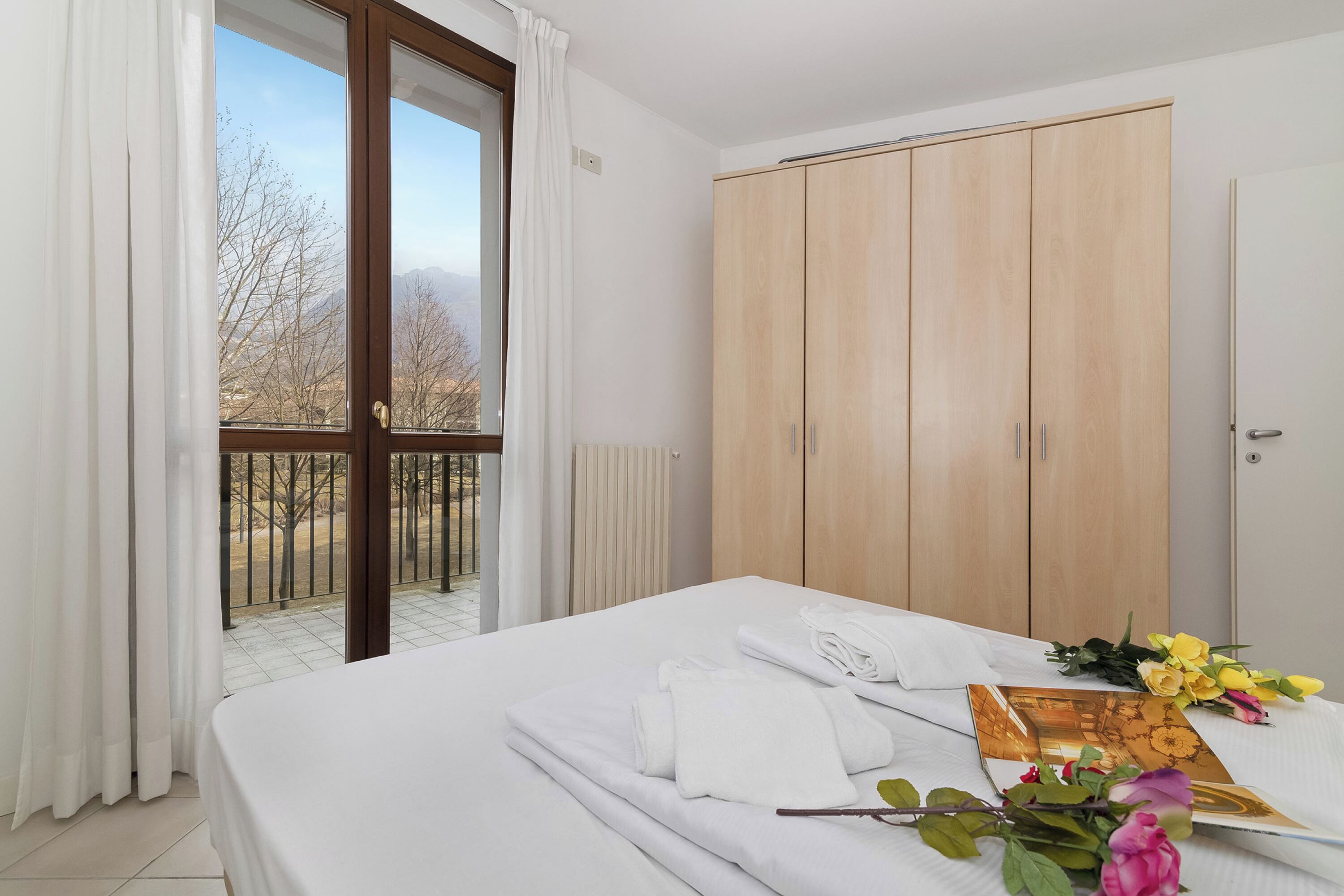 A hotel room with a bed, a wooden wardrobe, a balcony with a view of trees and mountains, and a table with towels and flowers.