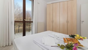 A hotel room with a bed, a wooden wardrobe, a balcony with a view of trees and mountains, and a table with towels and flowers. - Porto Letizia Tchaikovsky 7, Porlezza, Italy (Porlezza)