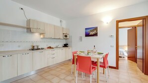 A modern kitchen with light wood cabinets, a dining table with red chairs, and a view into a bedroom through an open door. - Porto Letizia Roma 5, Porlezza, Italy (Porlezza)