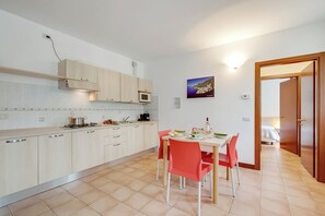 A modern kitchen with light wood cabinets, a dining table with red chairs, and a view into a bedroom through an open door.